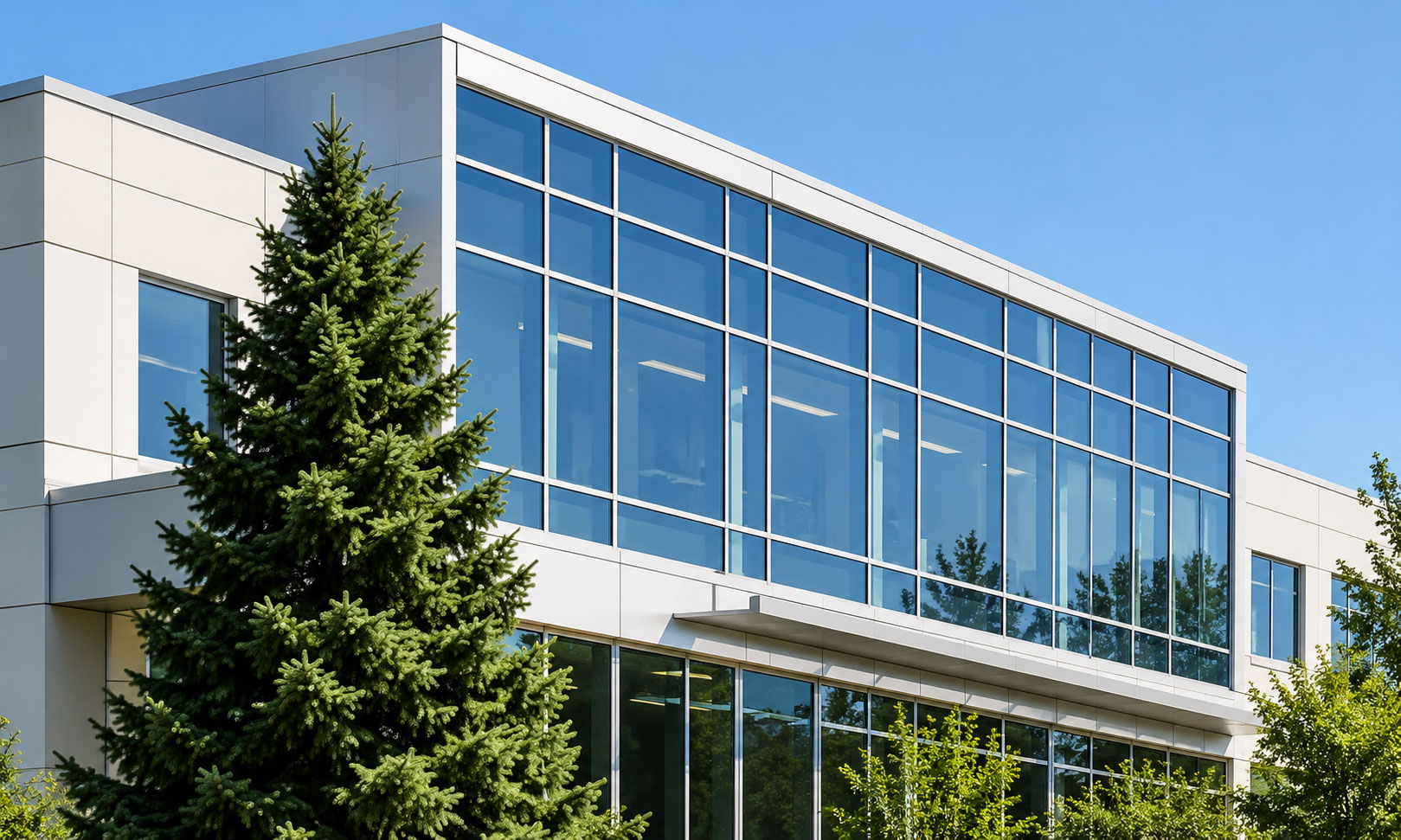 Contemporary office building with large glass windows reflecting blue sky and surrounding trees, featuring light-colored panel walls and a sleek rectangular design.