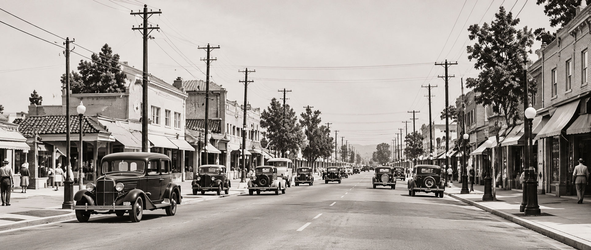 Black‑and‑white photograph of a busy town street from the early 1900s, showing vintage cars, pedestrians, storefronts with awnings, and utility poles lining both sides of the road.